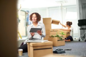 A business owner sits amongst her moving in boxes setting up VOIP, waiting for new desks. She is using a digital tablet. In the background her assistant is checking the files