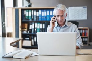 Mature business man at an office desk looking at a laptop while calling his managed IT team for help 