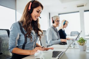 Three women smiling and working an IT help desk with headsets and laptops